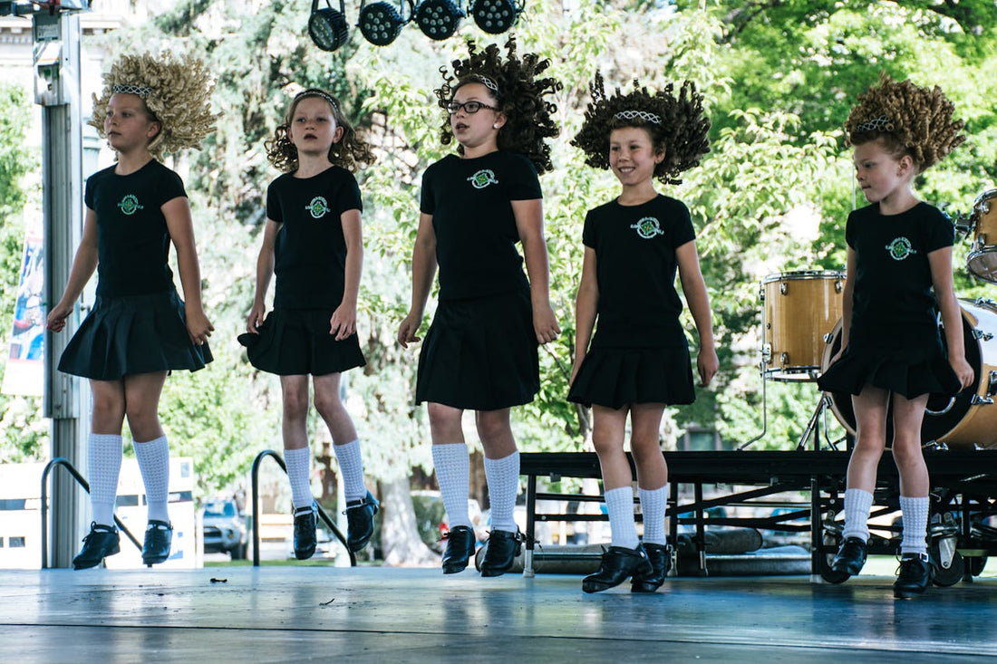 Group of young girls performing traditional Irish dance on stage, wearing black outfits, white socks, and curly wigs