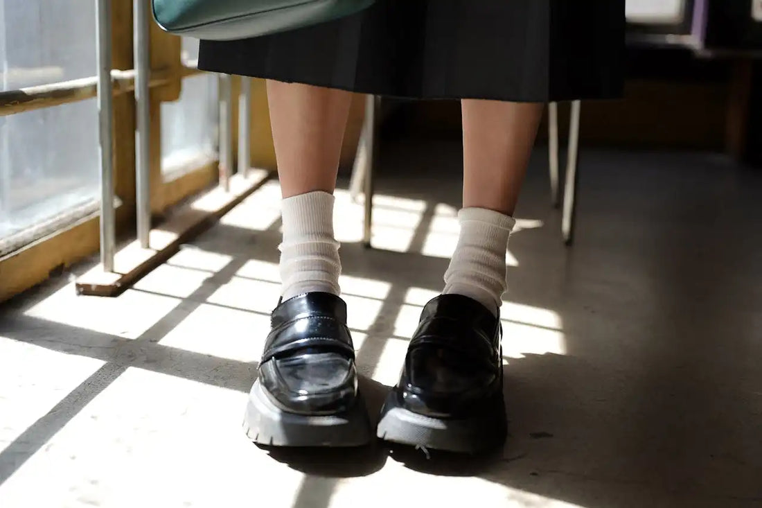 Close-up of a person wearing black leather loafers with white socks, sitting indoors with sunlight casting shadows on the floor
