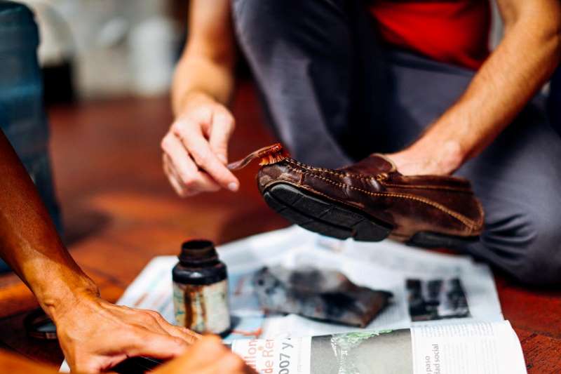 Person polishing a brown leather shoe with a brush, using shoe polish on a newspaper-covered floor